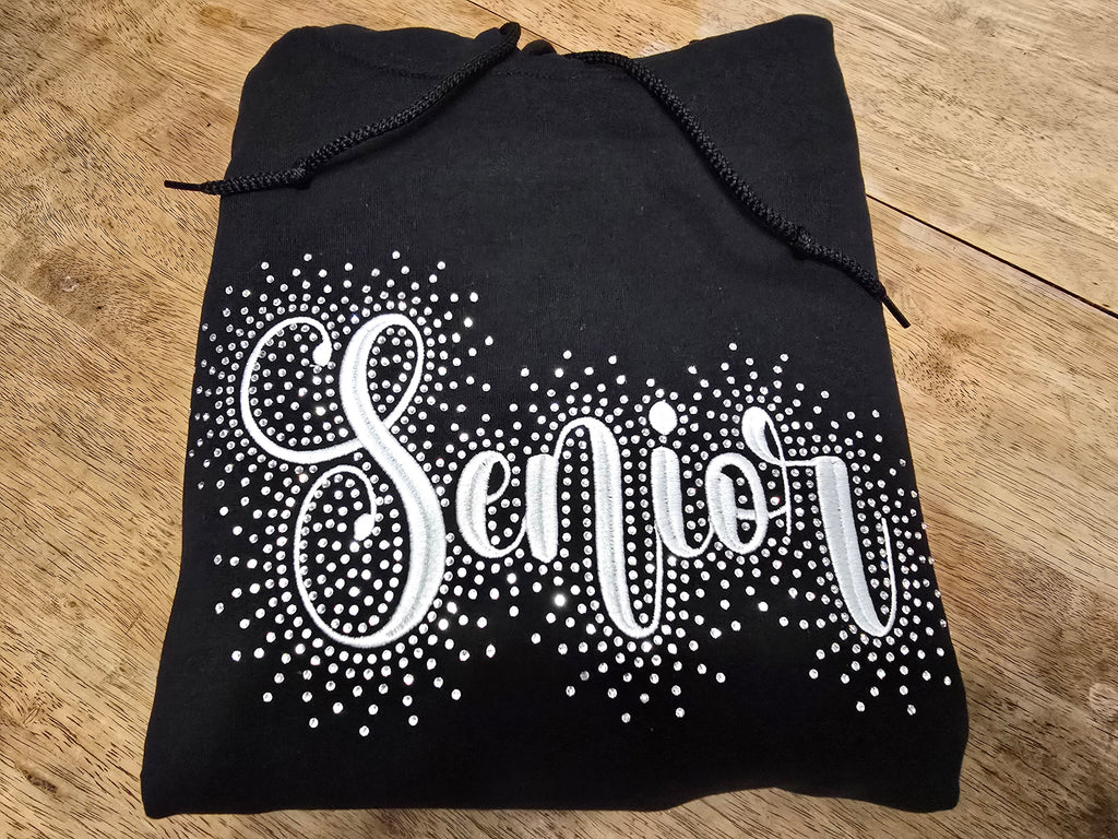 Black hoodie with 'Senior' embroidered on it with rhinestones on a wooden table.